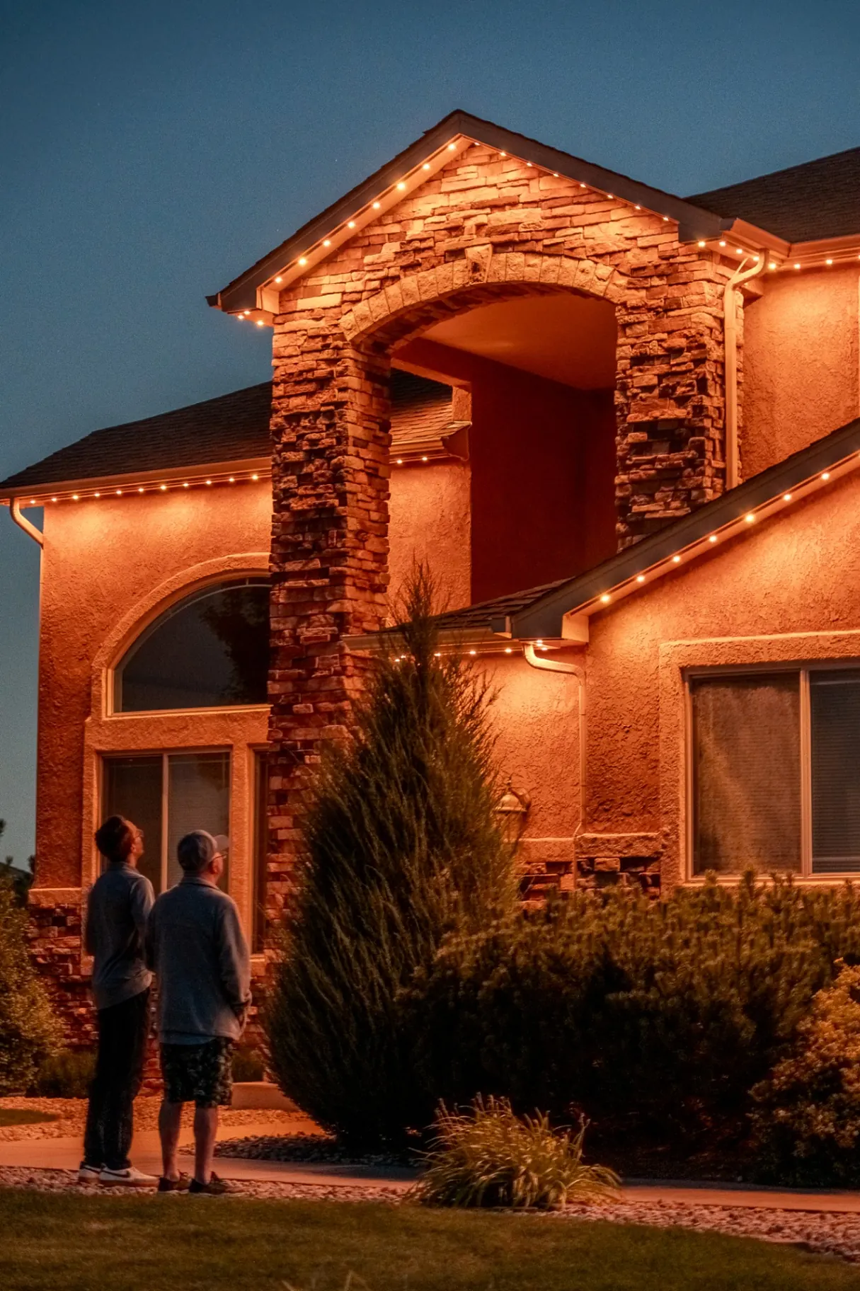 Warm white roofline lighting on a stone-front home with homeowners outside